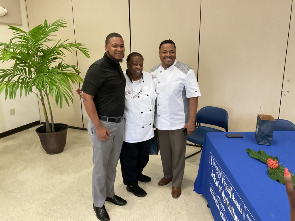 Three men stand together, smiling for the camera at an indoor event with a potted plant nearby.